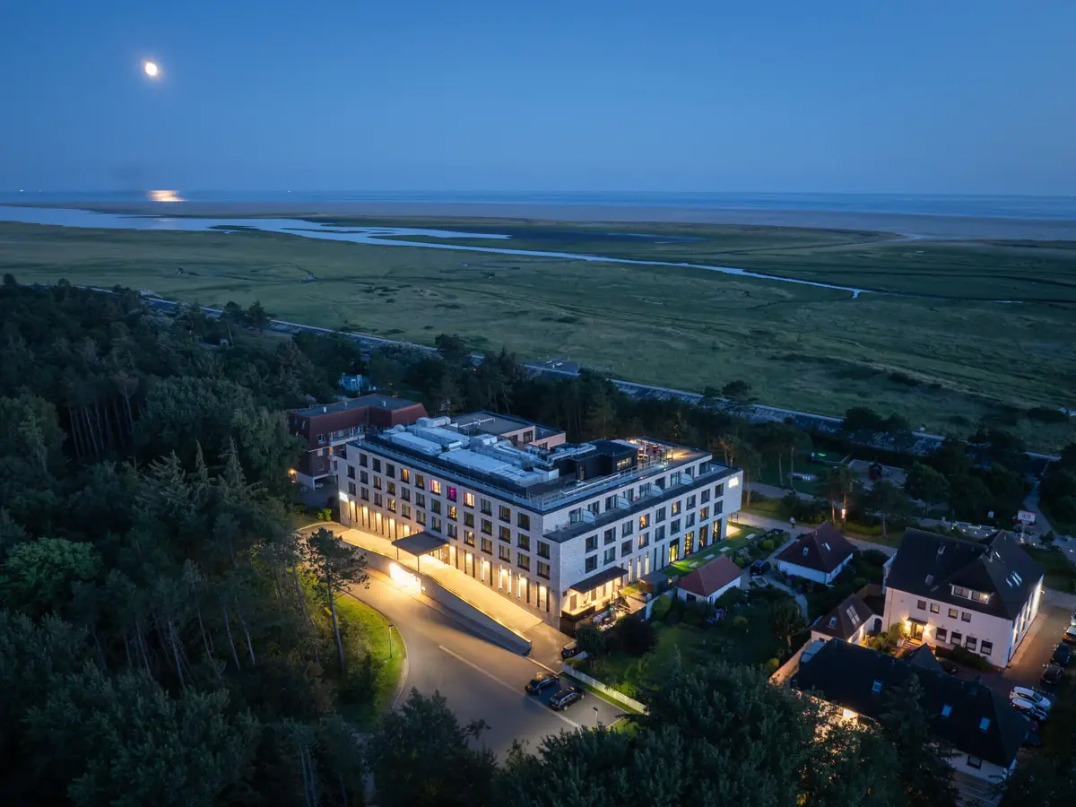 Urban Nature Hotel St. Peter-Ording Die Außenansicht vom Urban Nature Hotel St. Peter-Ording bei Dämmerung mit vielen Bäumen drum herum und Blick auf die Salzwiesen und der Nordsee im Hintergrund.