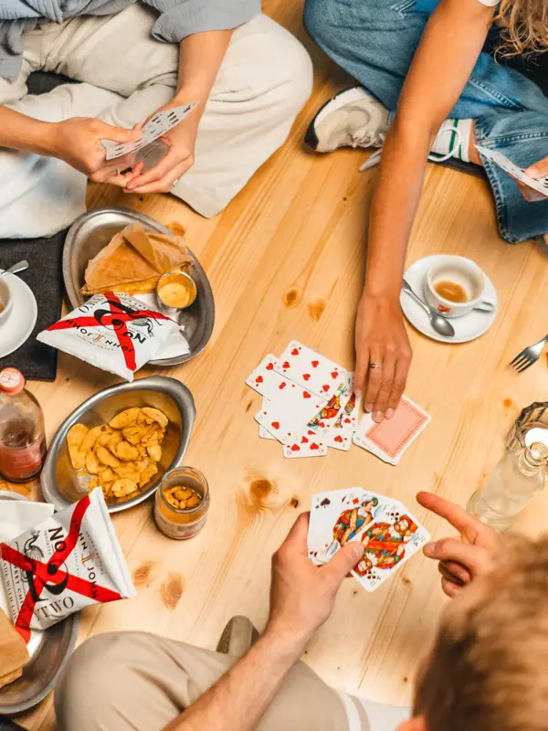 Drifters Hang Out Food Eine Gruppe von Menschen sitzt auf einem hölzernen Untergrund und spielt ein Kartenspiel während dazwischen mehrere Drinks und Snacks wie Chips stehen.