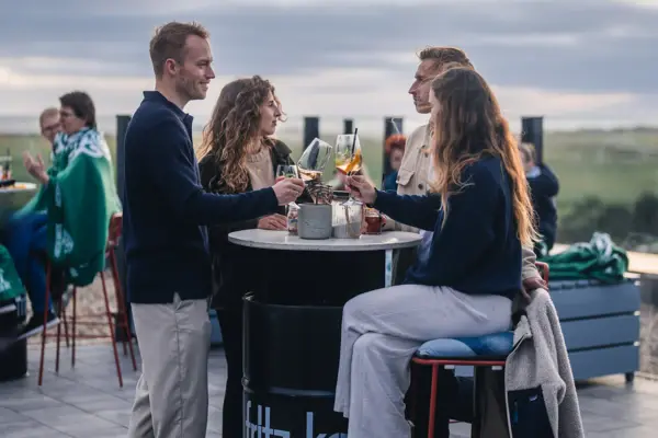Eine Gruppe von Menschen steht an einem Stehtisch auf einer Dachterrasse in St. Peter-Ording und stößt mit Getränken an.