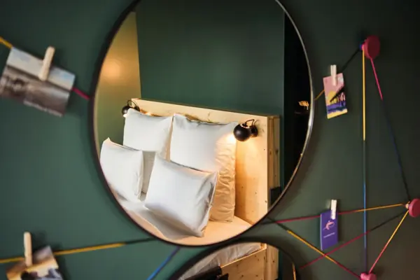 In einem Spiegel an einer grünen Wand ist ein gemütliches Bett zu sehen mit weißer Bettwäsche und vier Kissen.