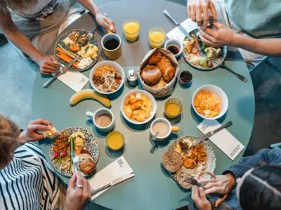 Ein runder Frühstückstisch von oben fotografiert, an dem vier Menschen sitzen und Brötchen, Obst und weitere Speisen essen.