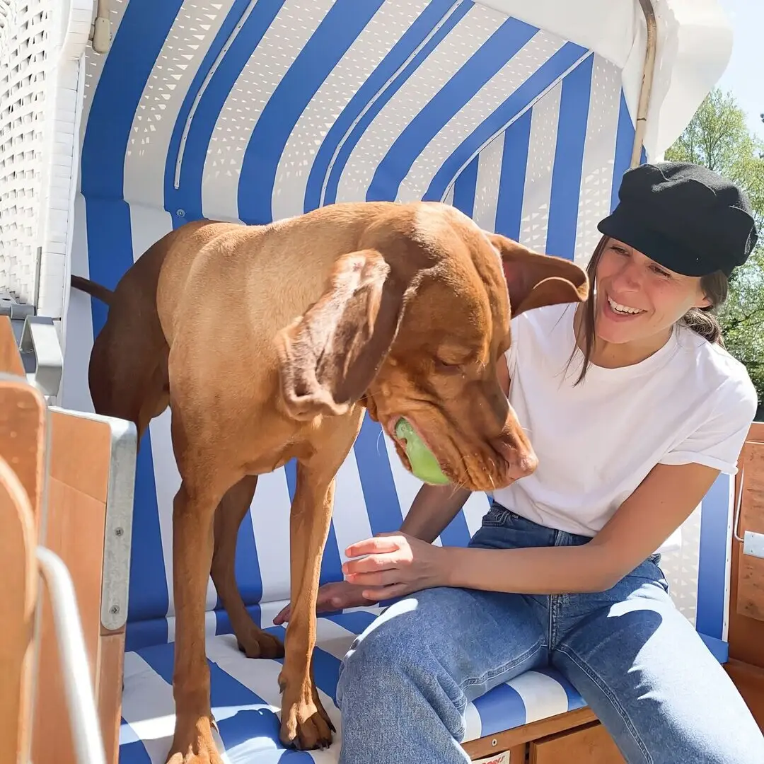 Eine Frau sitzt mit einem großen braunen Hund in einem Strandkorb und lächelt.