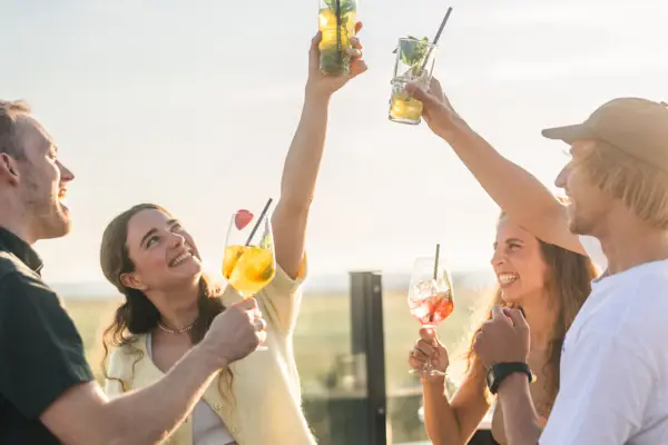 Die Rooftopbar Scraper's Club im Urban Nature Hotel St. Peter Ording mit 4 Personen, die bei Sonnenschein mit Drinks anstoßen und die Drinks hochhalten..