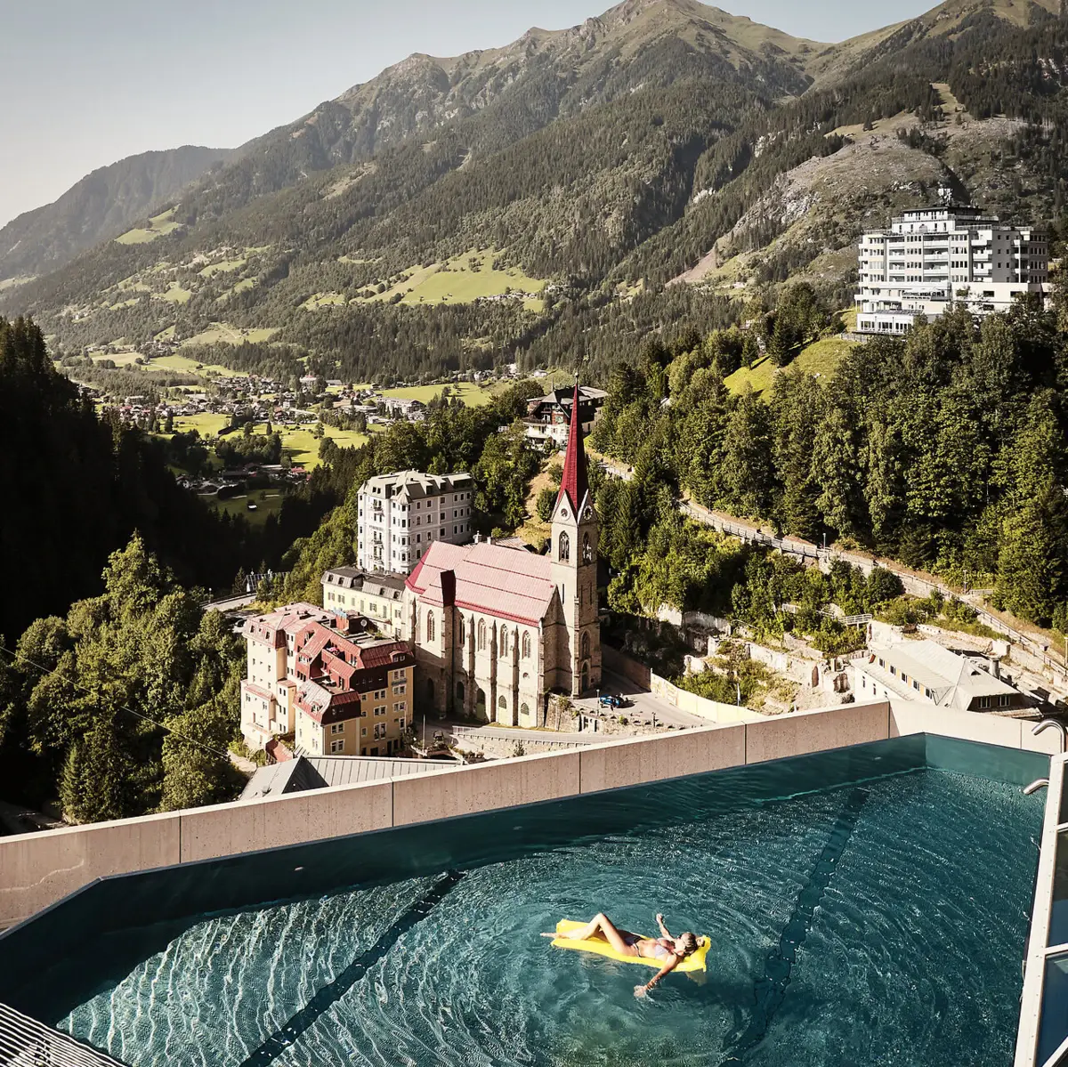 Infinity Pool mit Ausblick auf Bad Gastein und die Alpenlandschaft – Frau entspannt auf gelber Luftmatratze mit Blick auf Kirche und historische Gebäude