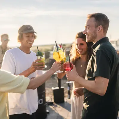 Eine Gruppe von Menschen steht auf der Rooftop Bar Scraper's Club St. Peter-Ording mit Getränken in der Hand und mit Blick auf die Salzwiesen und das Meer.