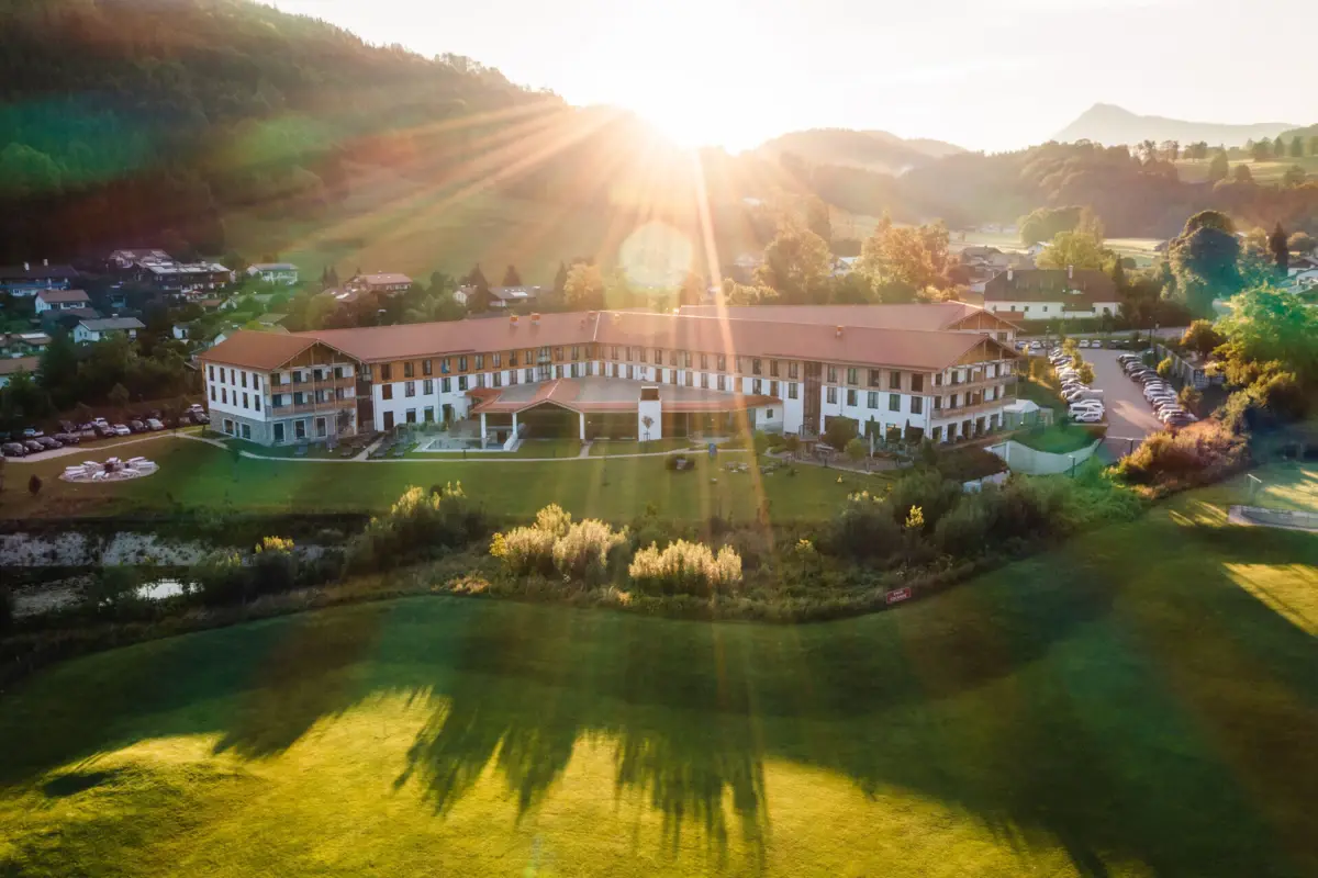 Luftaufnahme eines großen Hotels mit weitläufigem Garten in der Alpenlandschaft, Sonnenstrahlen strahlen über die Hügel und den Morgenhimmel
