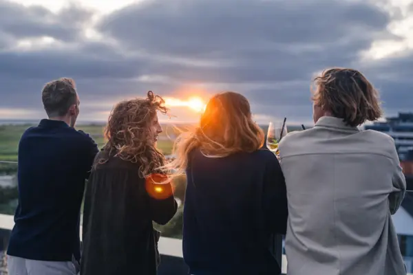 Eine Gruppe von Menschen steht auf der Rooftop Bar Scraper's Club St. Peter-Ording und schaut den Sonnenuntergang an.