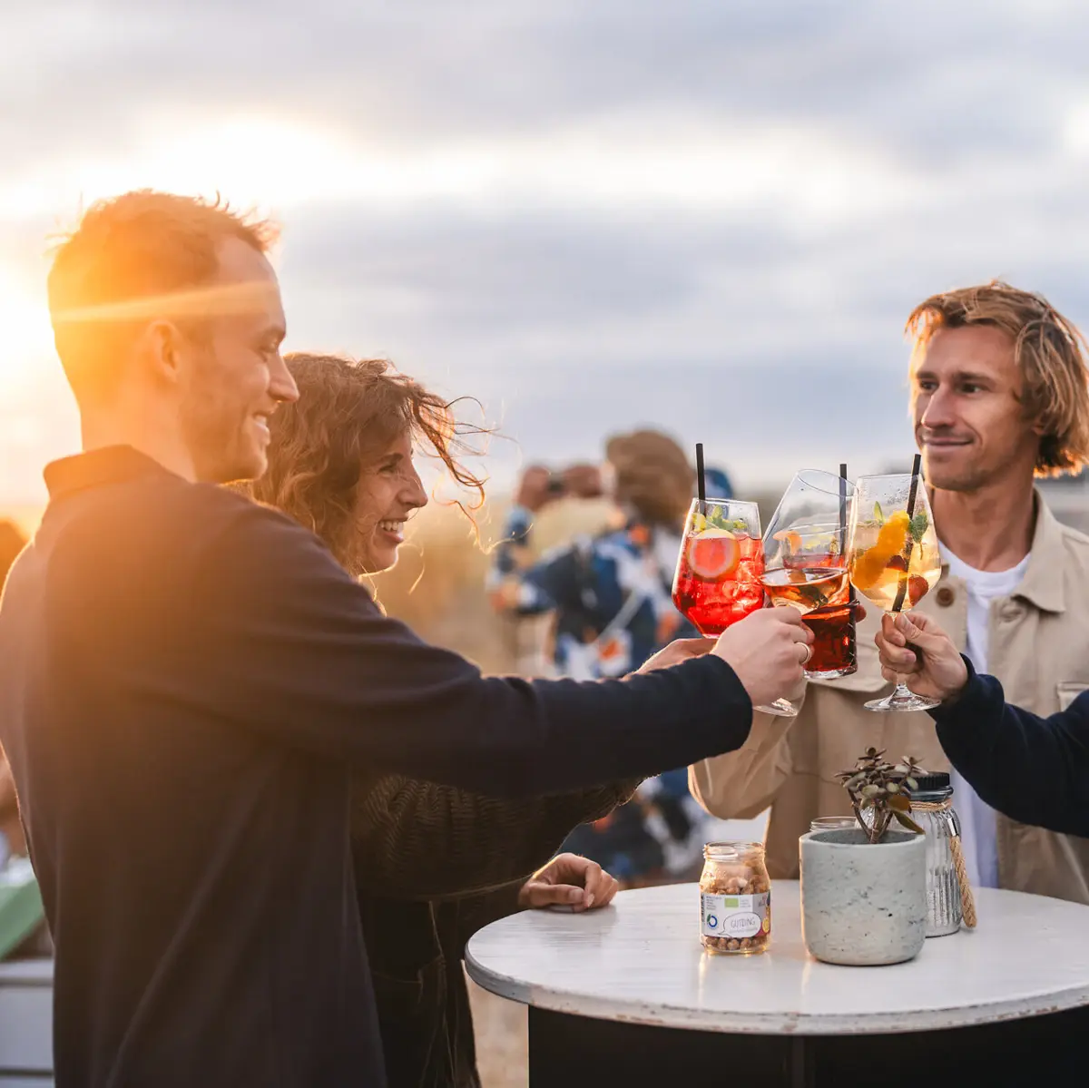 Eine Gruppe von Menschen steht auf der Rooftop Bar Scraper's Club St. Peter-Ording mit Getränken in der Hand und stößt beim Sonnenuntergang an.