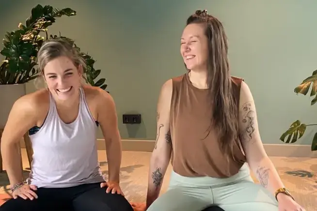 Meike & Agnes Zwei Frauen sitzen lächelnd auf einer Yogamatte im Schneidersitz.