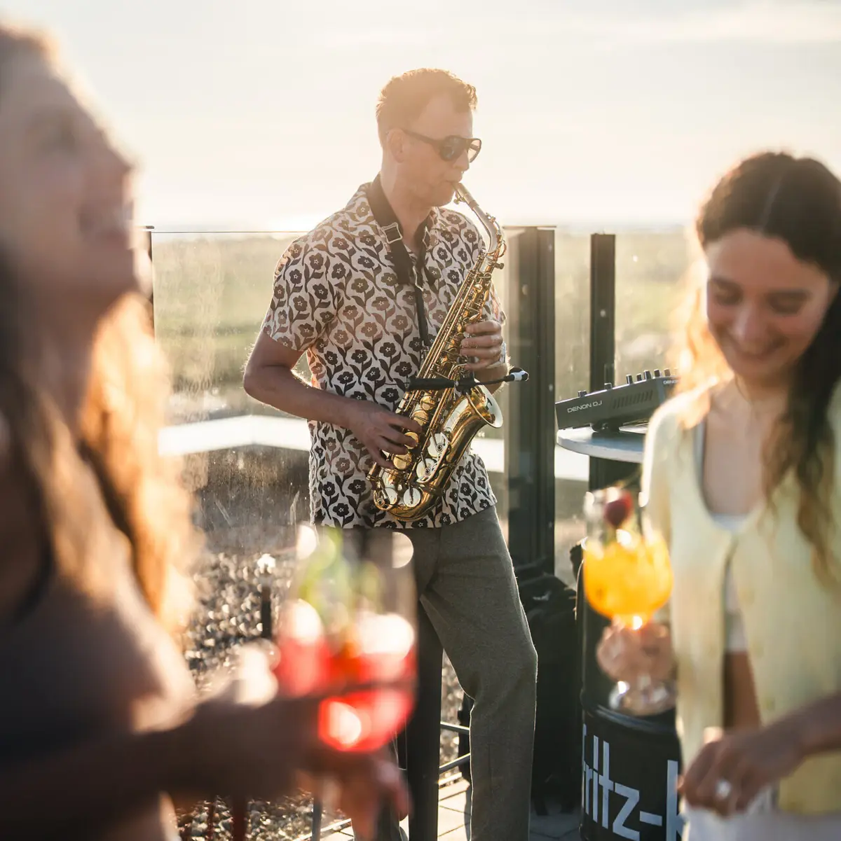 Ein Saxophonist spielt auf einer Dachterrasse während Leute mit Drinks in der Hand tanzen.