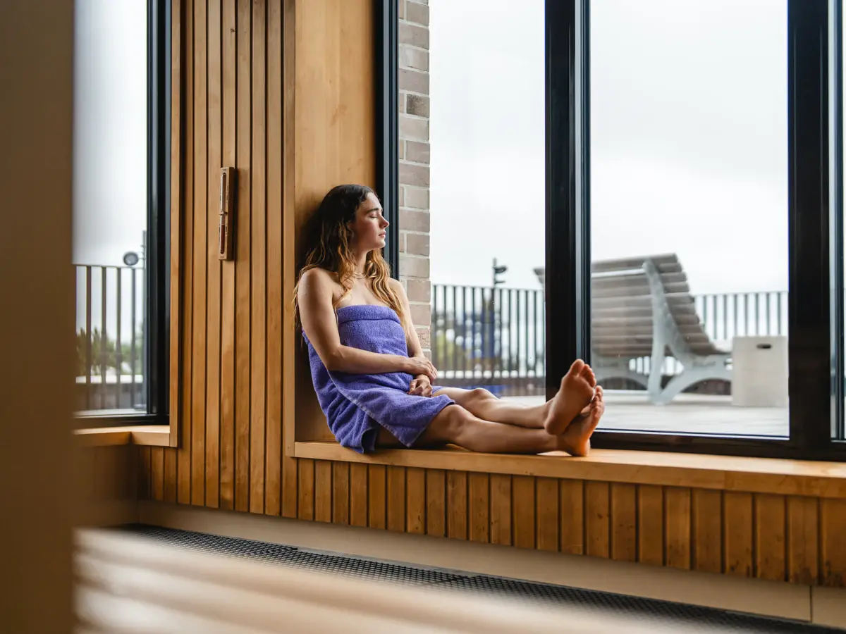 Sauna St. Peter-Ording Eine Frau sitzt auf an einem Fenster in einer Sauna und hat ein lilanes Handtuch um ihrem Körper.