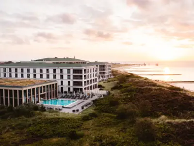 Modernes Hotel mit Außenpool direkt an der Küste, umgeben von Dünenlandschaft, Sonnenaufgang über der Nordsee im Hintergrund