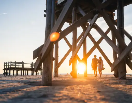 Zwei Menschen laufen Arm in Arm beim Sonnenuntergang am Strand von St. Peter Ording zwischen den Pfahlbauten entlang.