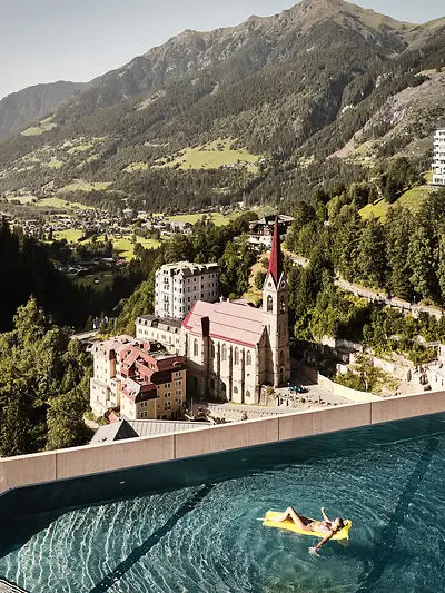 Infinity Pool mit Ausblick auf Bad Gastein und die Alpenlandschaft – Frau entspannt auf gelber Luftmatratze mit Blick auf Kirche und historische Gebäude