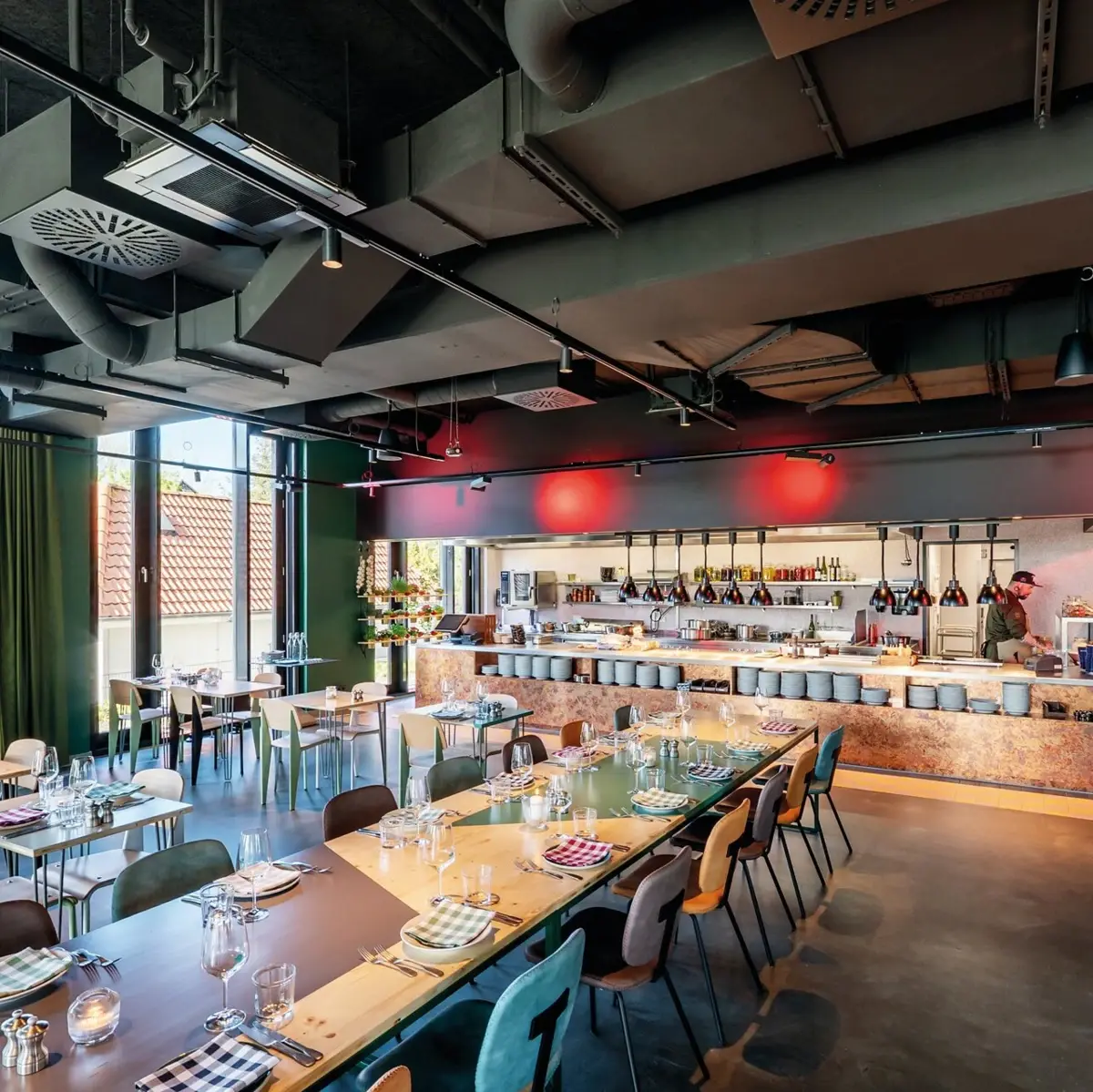 Ein Restaurant mit einer langen Tafel und mehreren gedeckten Tischen und einer offenen Küche im Hintergrund.