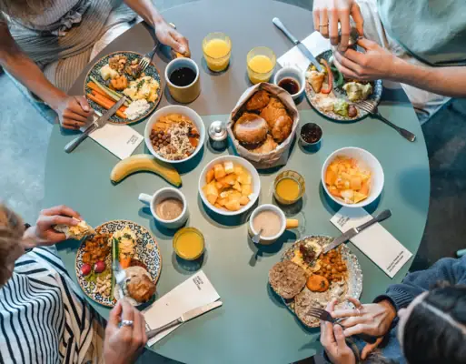 Ein runder Frühstückstisch von oben fotografiert, an dem vier Menschen sitzen und Brötchen, Obst und weitere Speisen essen.