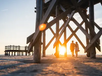Zwei Menschen laufen Arm in Arm beim Sonnenuntergang am Strand von St. Peter Ording zwischen den Pfahlbauten entlang.