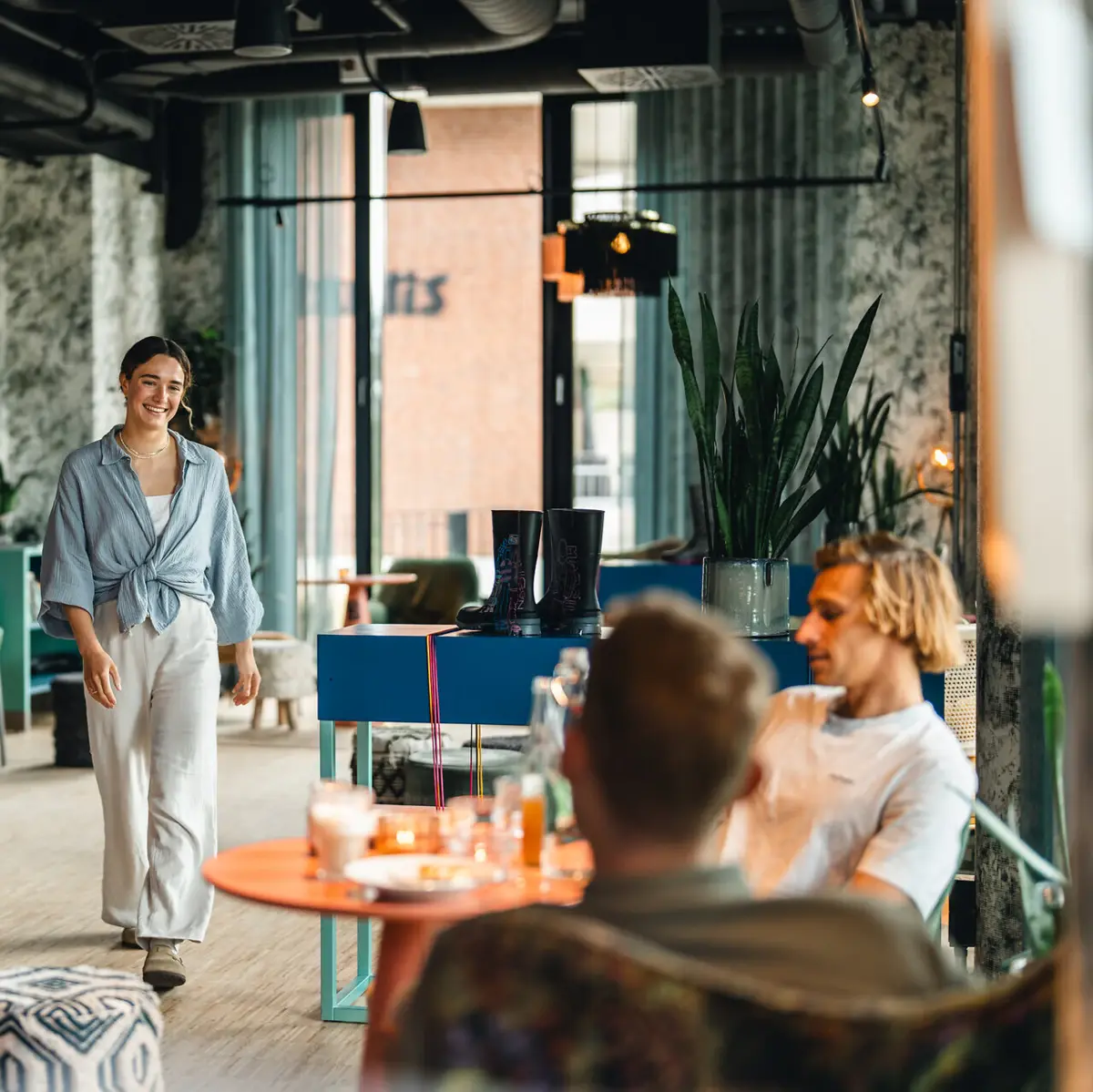 Eine Frau läuft in einer Bar auf einen Tisch zu, an dem schon zwei Männer sitzen und lächelt.