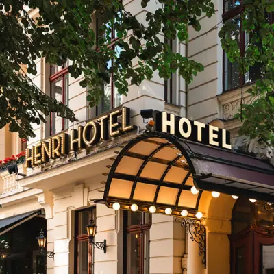 Eingang des HENRI Hotels in Berlin mit klassischem, eleganten Schriftzug und begrüntem Vordach, umgeben von alten Bäumen und historischer Fassade