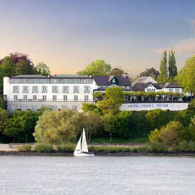 Hotel Louis C. Jacob mit Blick auf die Elbe, Segelboot im Vordergrund, umgeben von grüner Landschaft und Bäumen bei Sonnenuntergang
