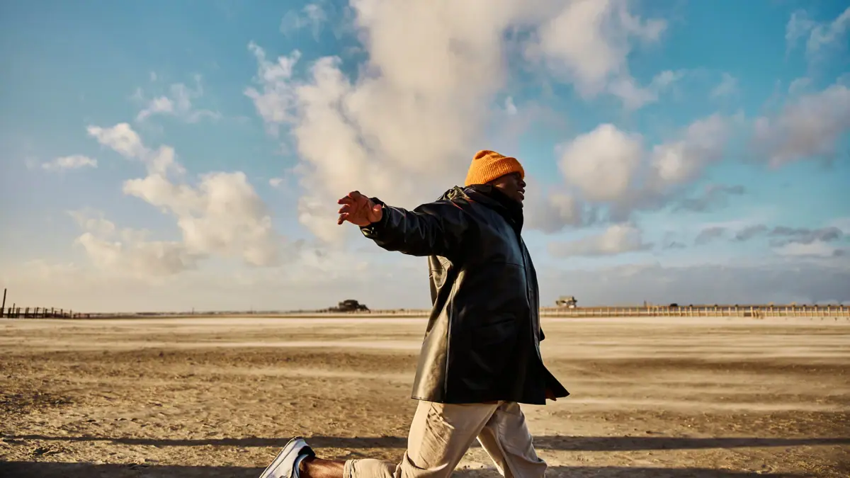 Ein Mann in schwarzem Mantel und orangefarbener Mütze am sandigen Strand.