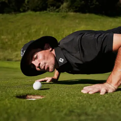 Golf Bad Gastein Ein Mann in Golf-Outfit und Hut stützt sich über dem Gras nach unten und schaut auf einen Golfball, der vor einem Loch liegt.
