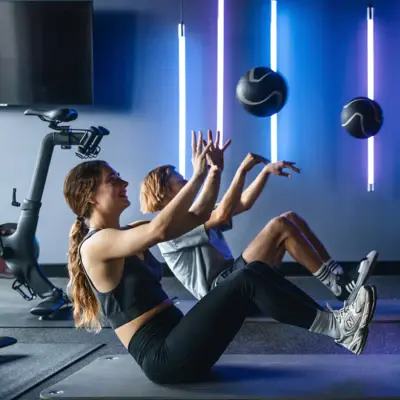 Zwei Personen sitzen auf einer Matte im Fitnessstudio, halten die Heine und den Oberkörper in der Luft und werfen dabei einen Ball.