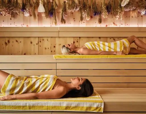 Zwei Frauen mit gelb-weiß gestreiften Handtüchern liegen entspannt in einer modernen Sauna mit Holzwänden und getrockneten Blumen an der Decke.