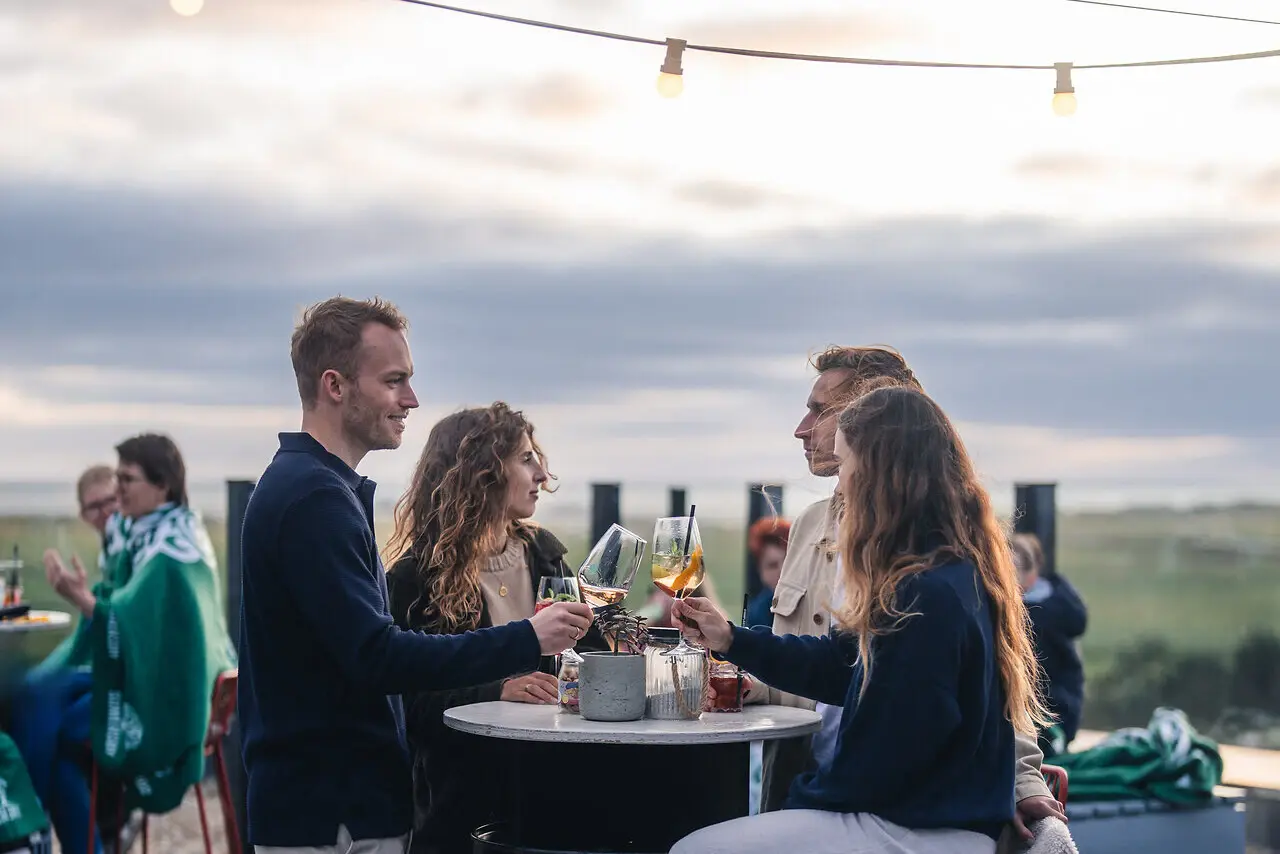 Rooftop Bar Scraper's Club St. Peter-Ording Eine Gruppe von Menschen steht auf der Rooftop Bar Scraper's Club St. Peter-Ording mit Getränken in der Hand und mit Blick auf die Salzwiesen und das Meer.