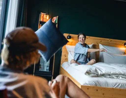 Eine Frau sitzt auf einem Bett in einem Zimmer im Urban Nature Hotel St. Peter-Ording und wirft ein Kissen auf die Kamera zu.