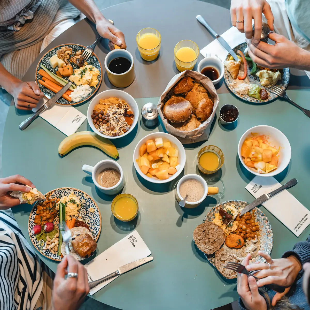 Ein runder Frühstückstisch von oben fotografiert, an dem vier Menschen sitzen und Brötchen, Obst und weitere Speisen essen.