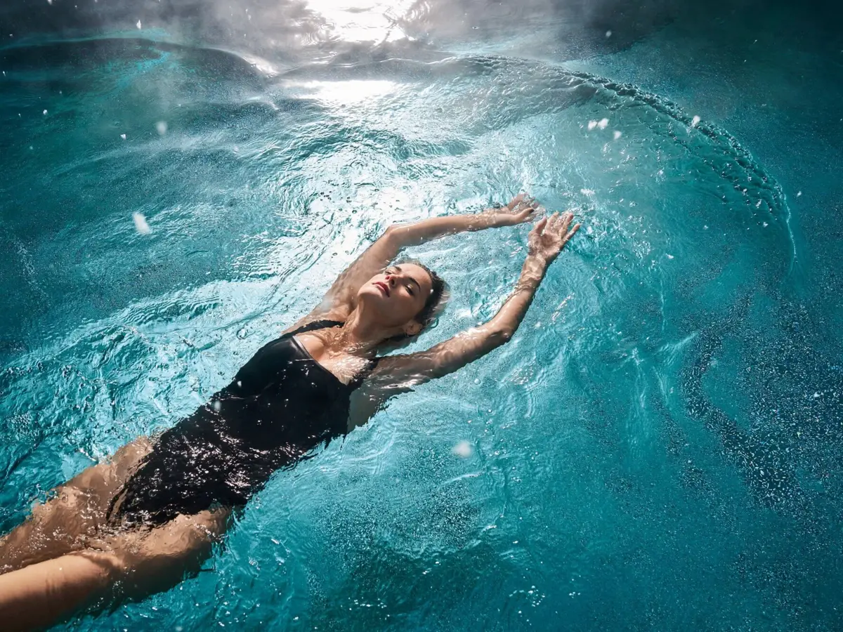 Alpine Swim Club Frau in schwarzem Badeanzug treibt entspannt auf dem Rücken in einem sonnendurchfluteten Pool.