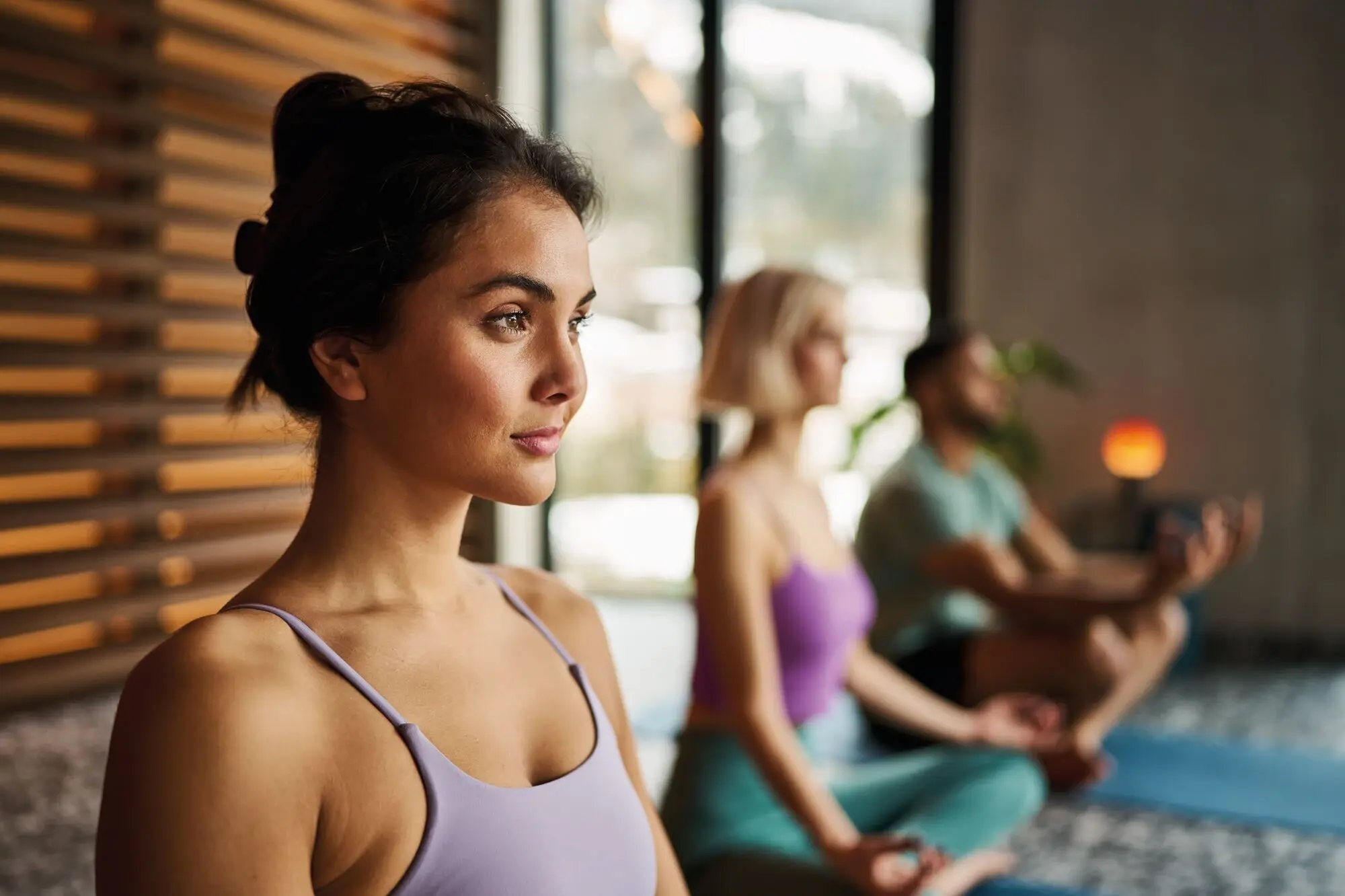 Yoga im Alpine Swim Club Junge Frau in Yoga-Pose mit geschlossenen Augen, im Vordergrund einer Meditationsgruppe in modernem, lichtdurchflutetem Raum