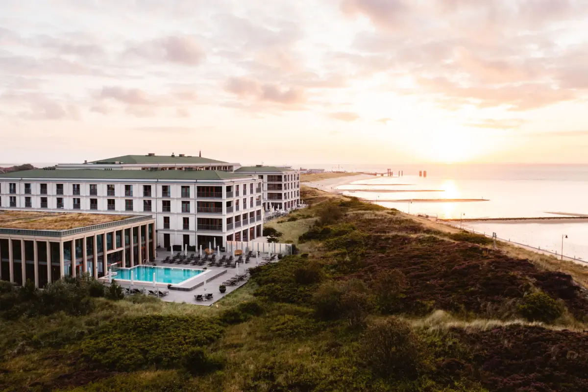 Modernes Hotel mit Außenpool direkt an der Küste, umgeben von Dünenlandschaft, Sonnenaufgang über der Nordsee im Hintergrund