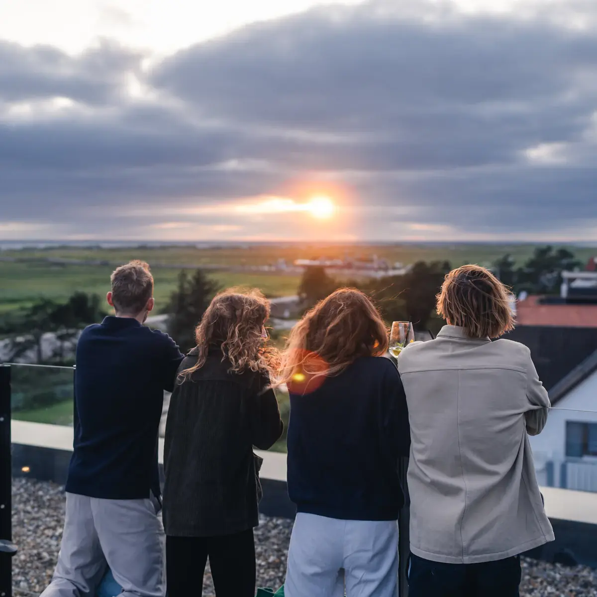 Eine Gruppe von Menschen steht auf der Rooftop Bar Scraper's Club St. Peter-Ording mit Blick auf die Salzwiesen und das Meer bei Sonnenuntergang.