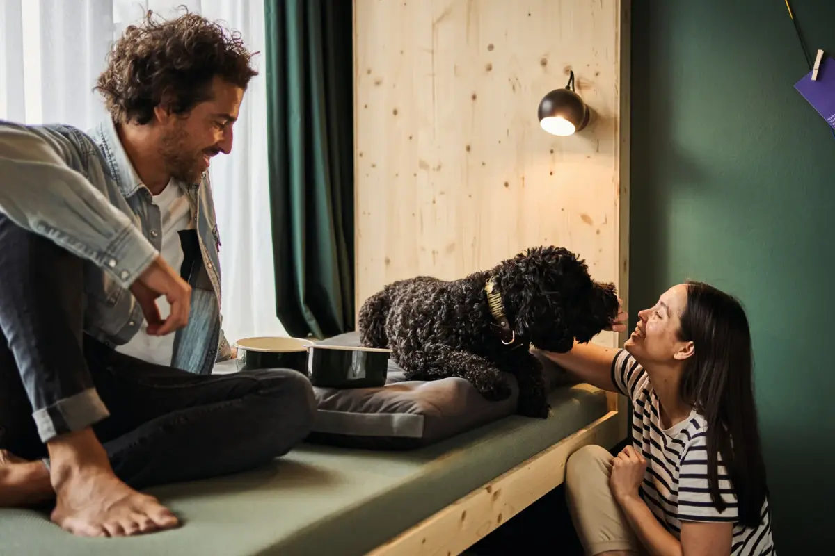 Ein Mann und eine Frau sitzen mit einem Hund auf einem Daybed in einem Hotelzimmer im Urban Nature Hotel St. Peter-Ording.