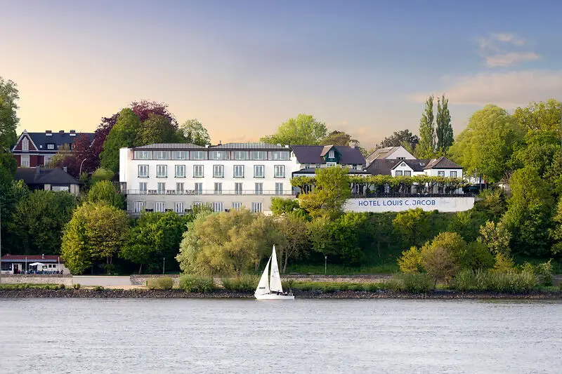 Hotel Louis C. Jacob mit Blick auf die Elbe, Segelboot im Vordergrund, umgeben von grüner Landschaft und Bäumen bei Sonnenuntergang