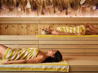 Zwei Frauen mit gelb-weiß gestreiften Handtüchern liegen entspannt in einer modernen Sauna mit Holzwänden und getrockneten Blumen an der Decke.
