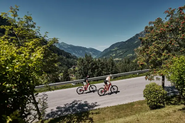Zwei Personen fahren mit Fahrrädern auf einer Straße, im Hintergrund sind Bäume und Berge zu sehen.