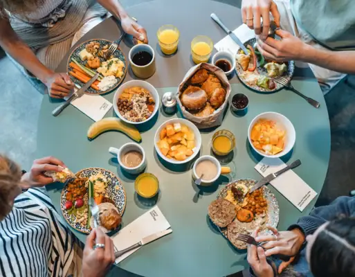 Ein runder Frühstückstisch von oben fotografiert, an dem vier Menschen sitzen und Brötchen, Obst und weitere Speisen essen.