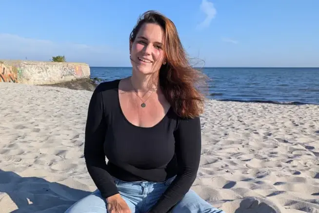 Yogalehrerin Ann-Katrin Feldkamp am Strand Die Yogalehrerin Ann-Katrin Feldkamp sitzt am Strand mit gekreuzten Beinen auf Sand und das Meer ist im Hintergrund zu sehen.