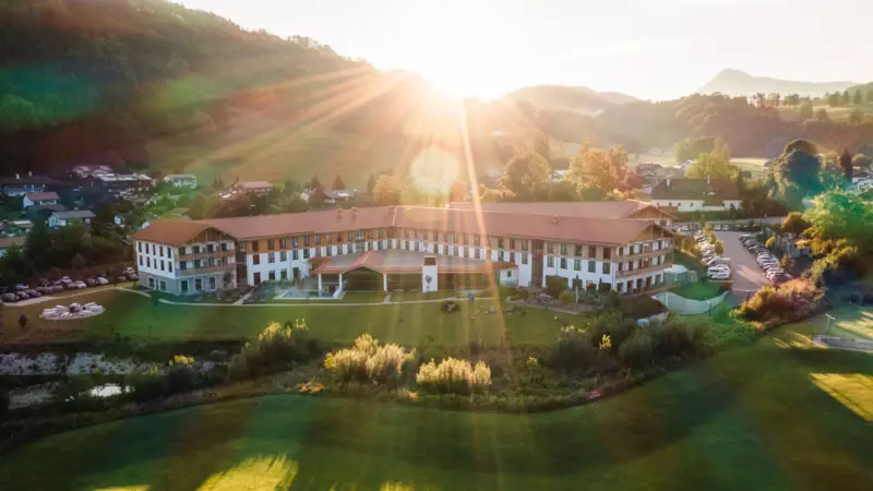 Luftaufnahme eines großen Hotels mit weitläufigem Garten in der Alpenlandschaft, Sonnenstrahlen strahlen über die Hügel und den Morgenhimmel