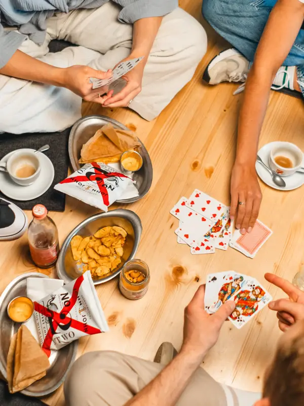 Mehrere Menschen sitzen im Schneidersitz auf einem Holzuntergrund und spielen zwischen Snacks ein Kartenspiel. 