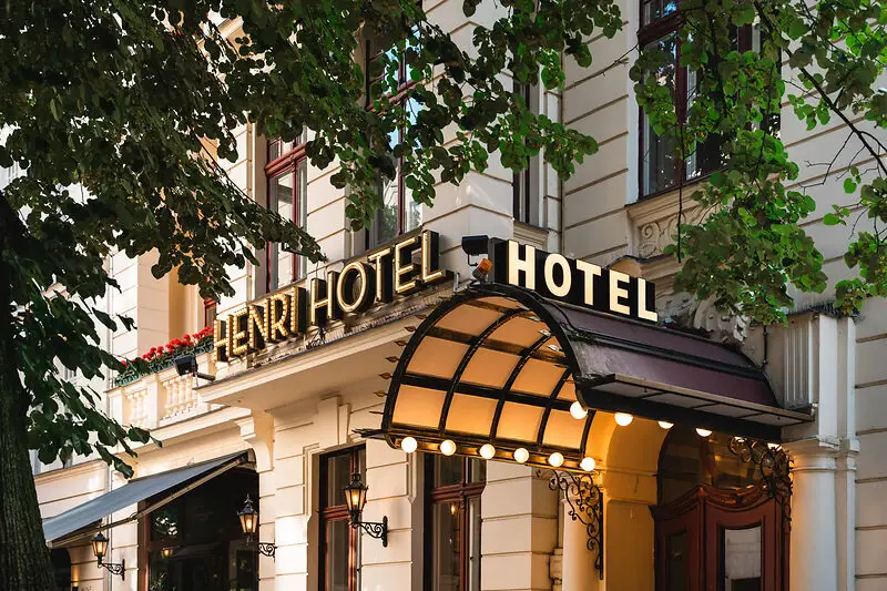 Eingang des HENRI Hotels in Berlin mit klassischem, eleganten Schriftzug und begrüntem Vordach, umgeben von alten Bäumen und historischer Fassade