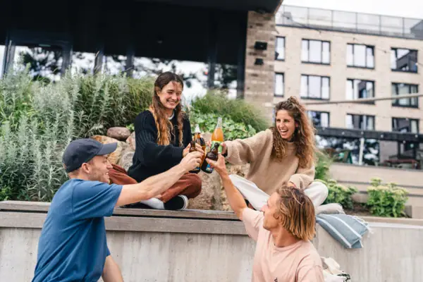 Eine Gruppe von Menschen sitzt auf der Plaza vom Urban Nature Hotel in St. Peter-Ording und stößt lachend mit Getränke-Flaschen an.