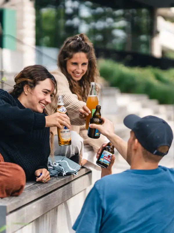 Eine Gruppe von Menschen sitzt auf einer Bank vor dem Urban Nature Hotel St. Peter-Ording und stoßt mit Bierflaschen an.