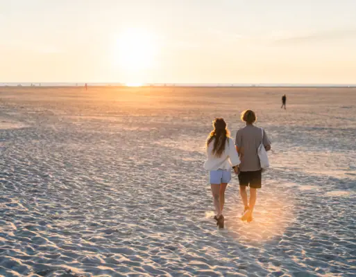 Ein Pärchen läuft Hand in Hand an einem Strand bei Sonnenuntergang lang.